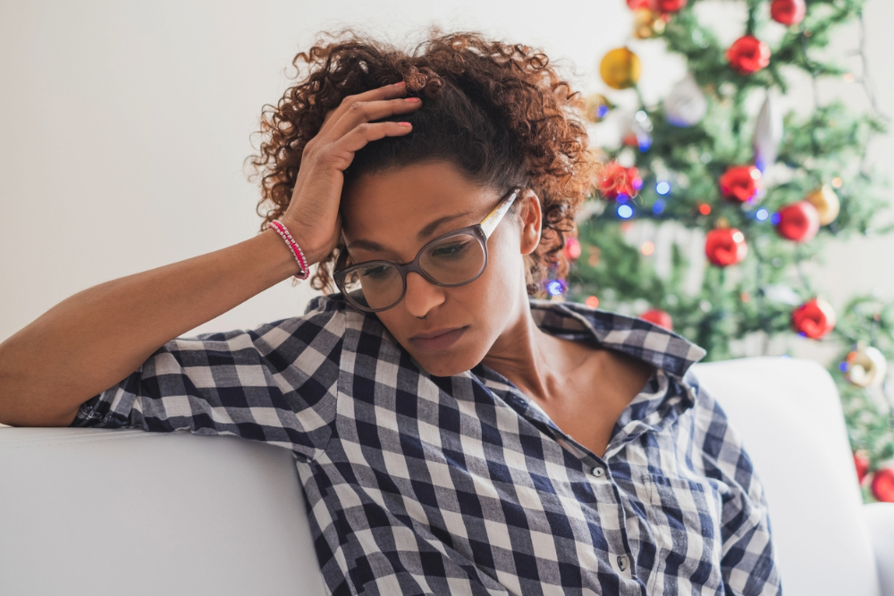 Black woman wondering how to avoid holiday depression on sofa in front of Christmas tree.