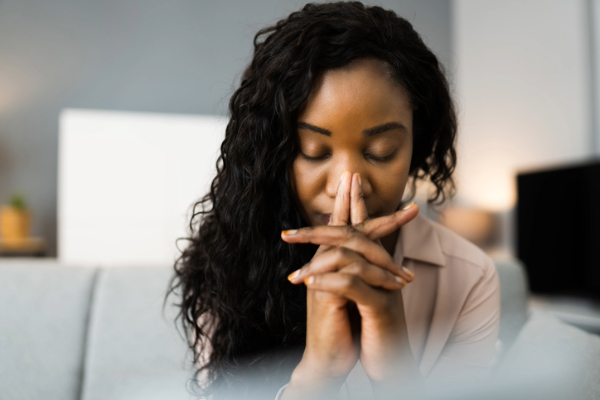 Worried young Black woman in prayer on sofa