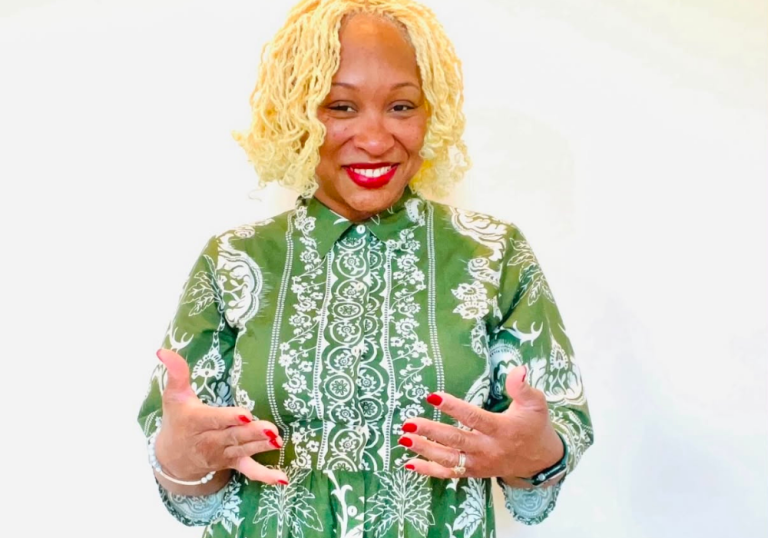 Smiling woman with short blonde curls in a green patterned blouse, gesturing with hands toward the camera.