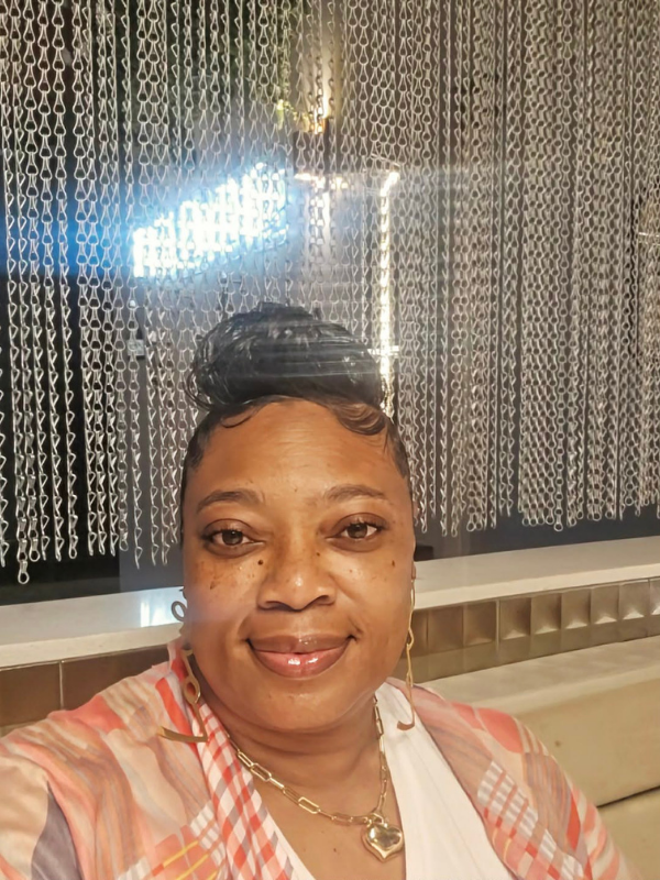 Smiling woman taking a selfie indoors, wearing a plaid jacket and gold heart necklace, with a beaded curtain backdrop and neon sign behind her.