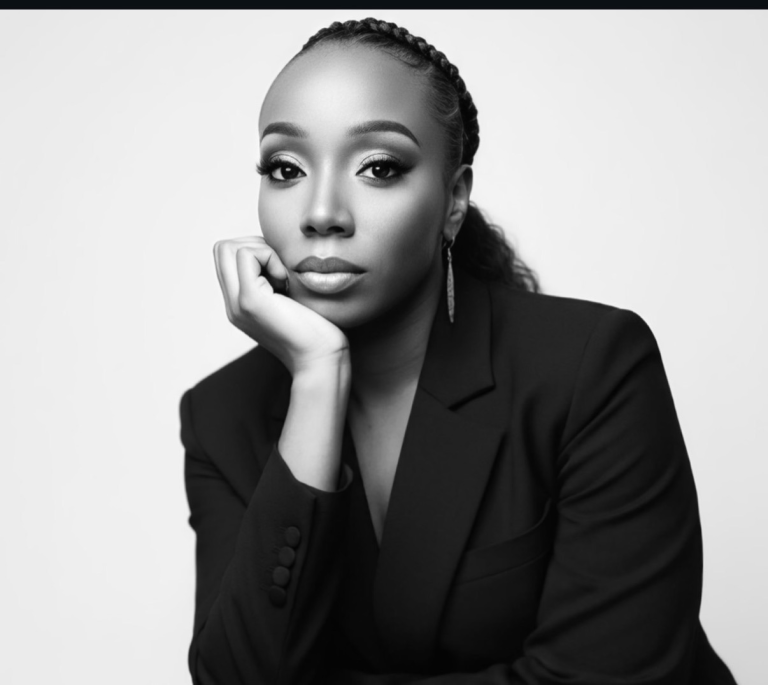 Black-and-white portrait of a woman in a blazer resting her chin on her hand, looking confidently at the camera, with braided hair.