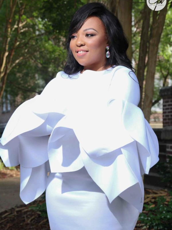 Woman in a white, oversized ruffled dress posing outdoors with a smile and drop earrings in a park setting.