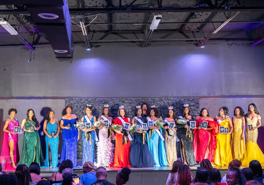 Group of pageant contestants in colorful gowns on stage, wearing crowns and holding plaques with bouquets, as an audience looks on.