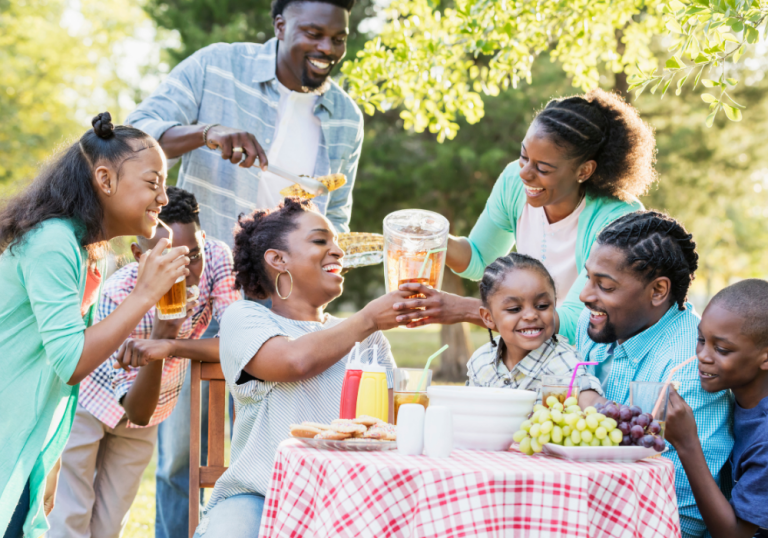 A diverse group of people enjoying an outdoor picnic, toasting drinks and smiling around a table with fruit and snacks on a sunny day.