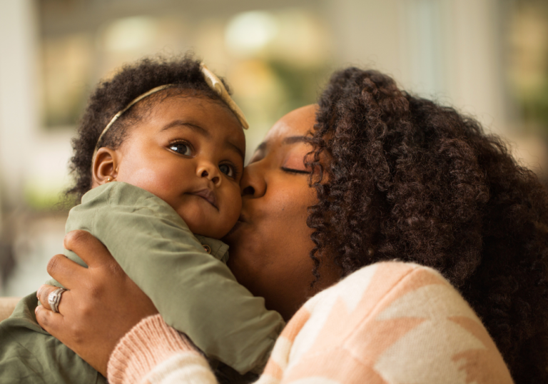 Mother lovingly kisses her baby on the cheek while holding them close indoors.