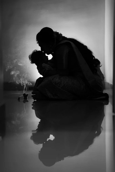 Silhouette of a mother holding her baby in a dim room, kneeling by a small incense burner with smoke, reflected on the floor.