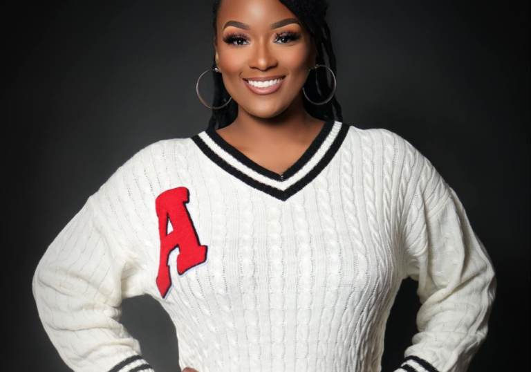 Smiling woman wearing a white cable-knit sweater with a red varsity-style 'A' patch on the chest.