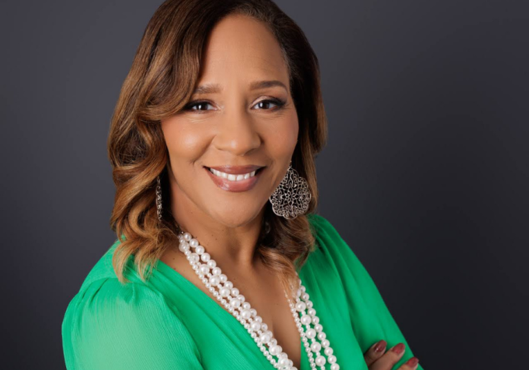 Professional headshot of a smiling woman with wavy brown hair, wearing a green blouse and layered pearl necklaces against a dark gray backdrop.