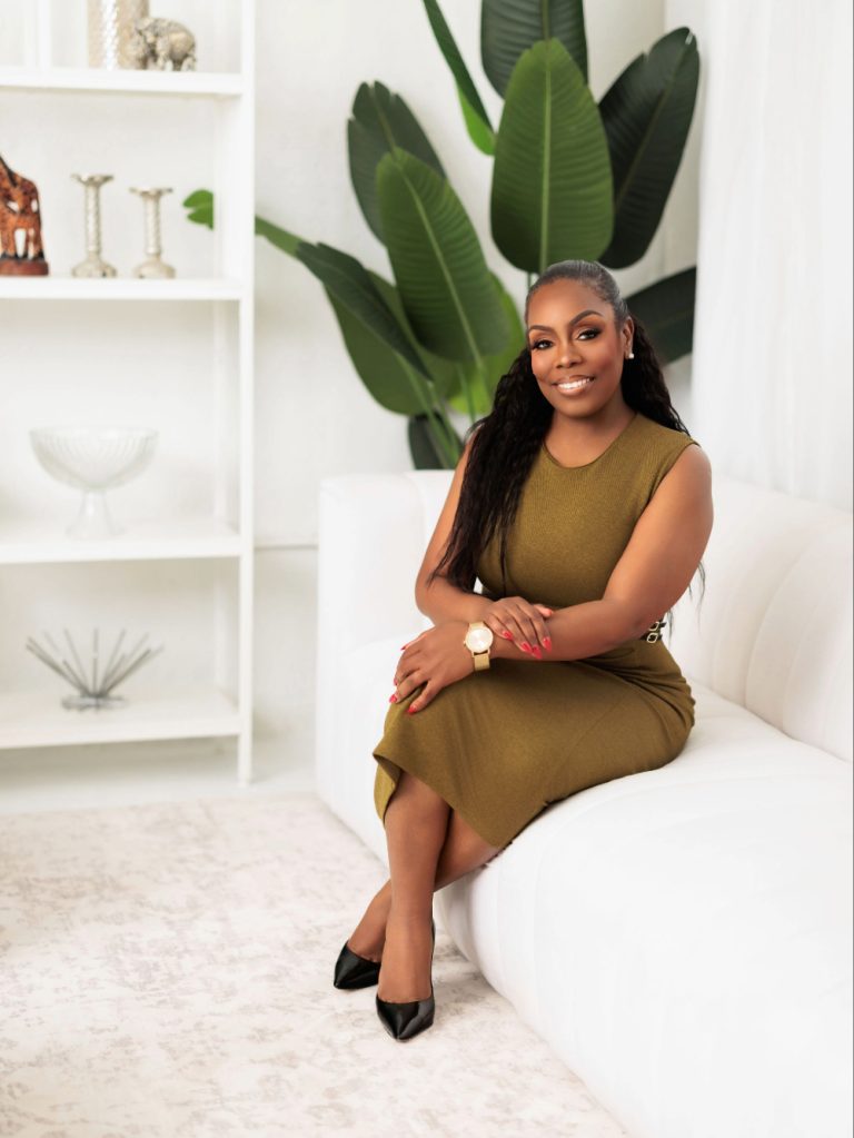 Smiling woman in an olive sleeveless dress sits on a white sofa in a bright living room with large green plants behind her.
