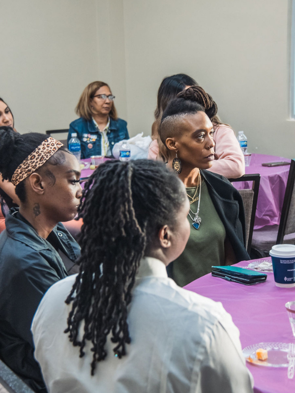 Group of women seated around a table with a purple tablecloth, attentively listening at a meeting or event in a conference room.