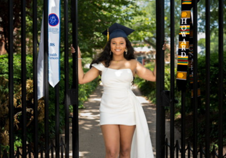 Graduate in a white off-shoulder dress and cap poses at an iron gate with campus banners, smiling at the camera.