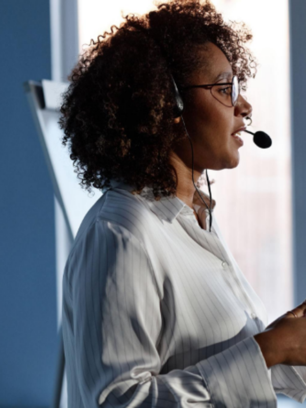 Profile of a woman with curly hair wearing a headset and microphone, speaking in a blue office setting.