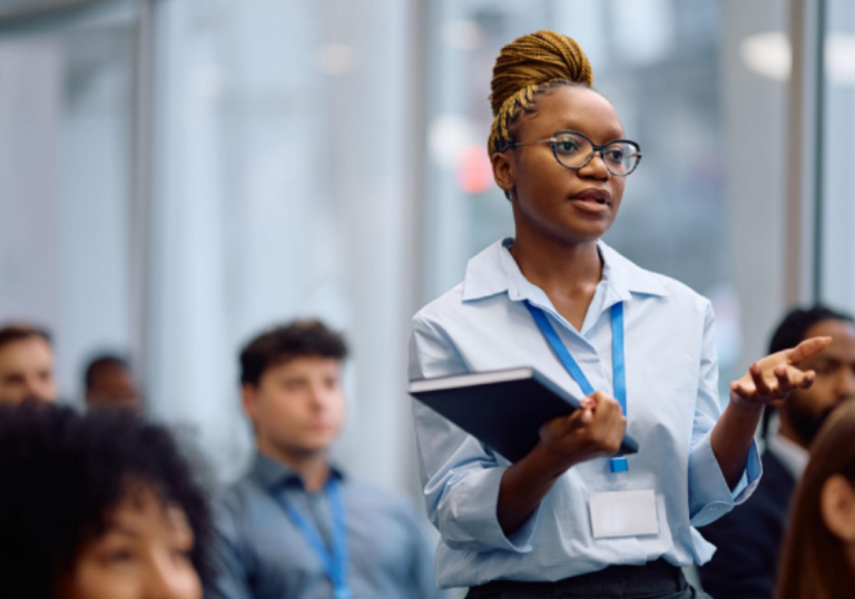 Professional woman presenting in a conference room, holding a notebook and gesturing as she speaks to an audience wearing conference lanyards