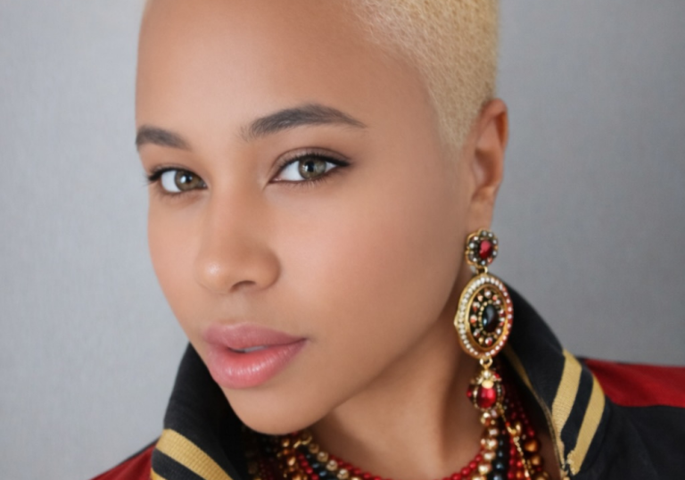 Close-up portrait of a person with platinum blonde hair, wearing large red-and-gold earrings and a beaded necklace, looking at the camera.