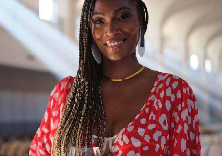 Smiling Black woman with long braided hair, wearing a red patterned blouse, large white earrings, and a gold necklace.