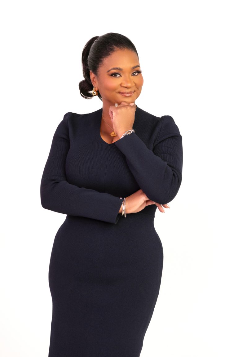 Tia Buckham-White Professional woman in a navy dress poses with her chin resting on her hand, smiling confidently against a white background.