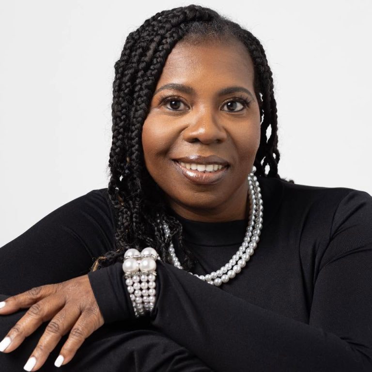 Close-up portrait of a smiling Black woman with braided hair, wearing a black top and layered pearl necklaces, resting her arms.