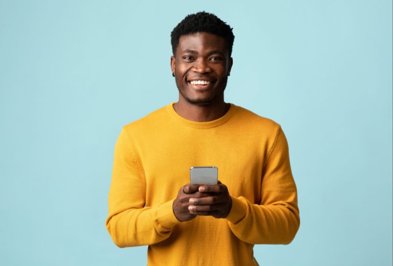 Smiling man in a mustard yellow sweater holding a smartphone toward the camera against a light blue background. (dating over 35)