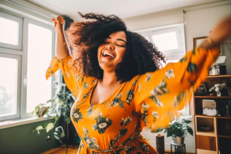 Woman with curly hair dancing joyfully in a bright room wearing an orange floral dress.