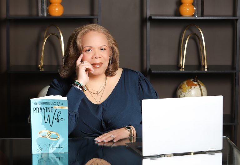 Woman in a navy blouse sits at a glass desk, resting her chin on her hand, with the book 'The Chronicles of a Praying Wife' standing upright in front of her.