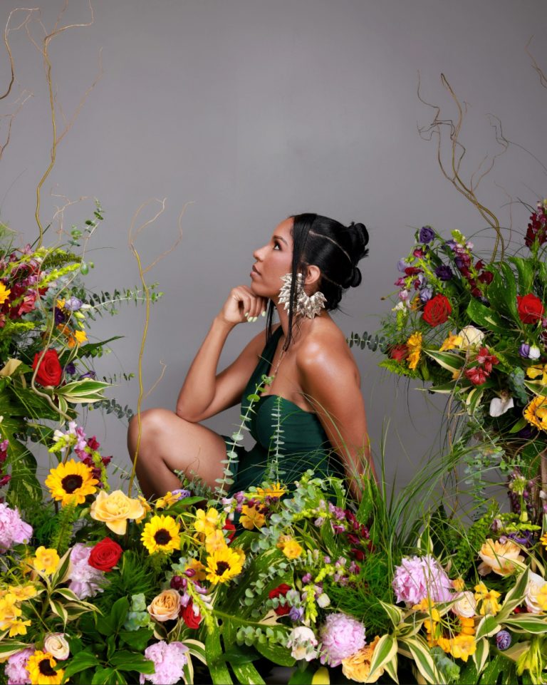 Woman in a dark green dress sits among a vivid bouquet of sunflowers, roses, and greenery, resting her chin on her hand in a contemplative pose.