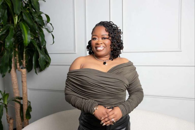 Smiling woman in an off-shoulder olive top posing indoors beside a tall plant and a light-colored sofa. She wears a necklace and rings, hands resting on her lap.