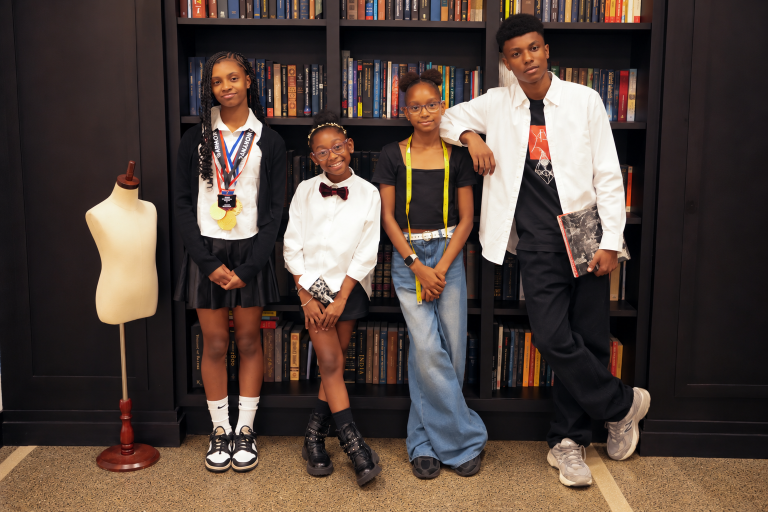 Image of four youth designers showcasing at Wearable Stories: Youth Community Couture, in front of a book shelf.